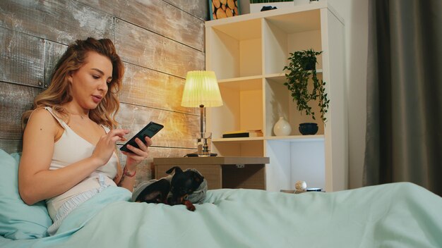 Junge Frau im Schlafanzug, die vor dem Schlafengehen auf ihrem Telefon scrollt. Glücklicher Hund.