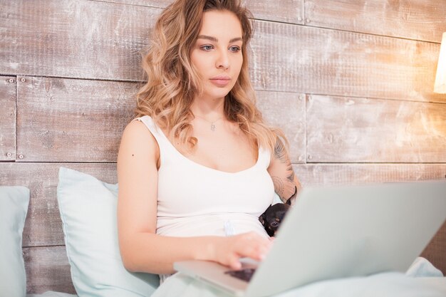 Junge Frau im Schlafanzug nachts in ihrem Bett, die zusammen mit ihrem kleinen Hund einen Film auf dem Laptop ansieht.