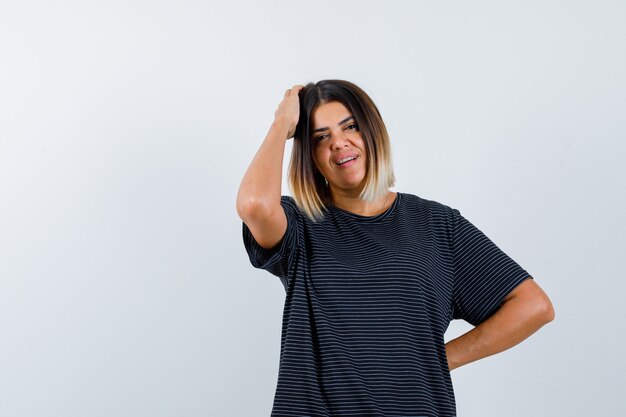 Junge Frau im schwarzen Kleid hält eine Hand auf Kopf, eine andere Hand auf Taille und sieht hübsch aus, Vorderansicht.