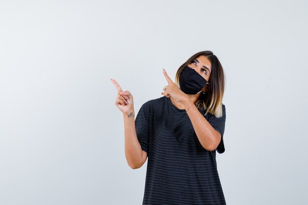 Junge Frau im schwarzen Kleid, schwarze Maske, die obere linke Seite mit Zeigefingern zeigt und fokussierte Vorderansicht schaut.