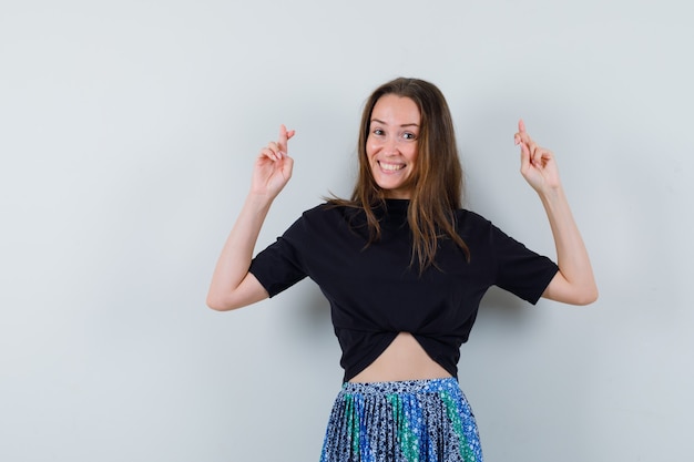 Junge Frau im schwarzen T-Shirt und im blauen Rock, die mit gekreuzten Fingern stehen und glücklich schauen