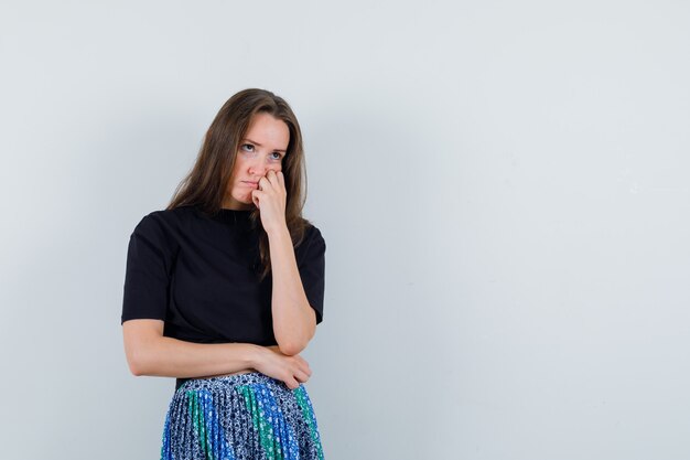 Junge Frau im schwarzen T-Shirt und im blauen Rock, die Wange auf Hand lehnt und über etwas nachdenkt und nachdenklich aussieht