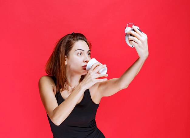 Junge Frau im schwarzen Unterhemd, die einen Wecker hält, während sie eine Tasse Tee trinkt