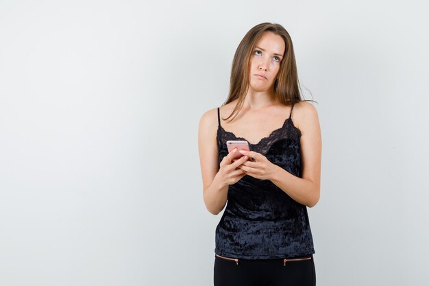 Junge Frau im schwarzen Unterhemd, Hosen, die Handy halten, während sie wegschauen und zögernd schauen