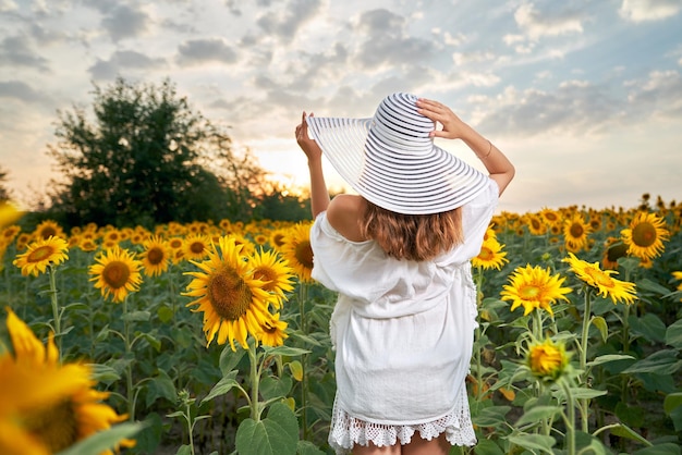 Junge Frau im Sommerhut, die mit Sonnenblumen auf dem Feld steht