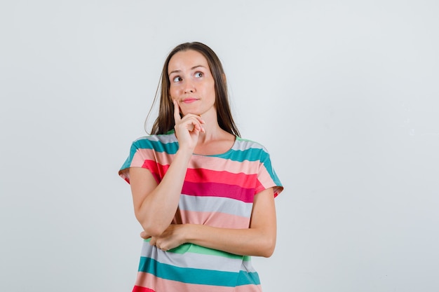 Junge Frau im T-Shirt, das Finger auf Wange hält und nachdenklich, Vorderansicht schaut.