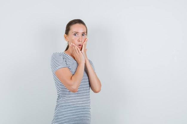 Junge Frau im T-Shirt, das Hände auf Wangen hält und ängstlich schaut, Vorderansicht.