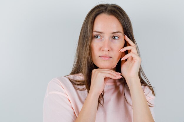 Junge Frau im T-Shirt, das Kinn auf Hand stützt, während Gesicht berührt und nachdenklich, Vorderansicht schaut.