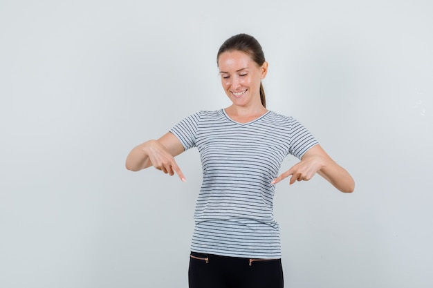 Junge Frau im T-Shirt, Hose zeigt nach unten und schaut fröhlich, Vorderansicht.