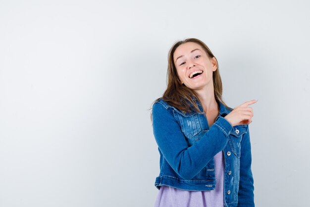 Junge Frau im T-Shirt, Jacke, die auf die obere rechte Ecke zeigt und fröhlich aussieht, Vorderansicht.