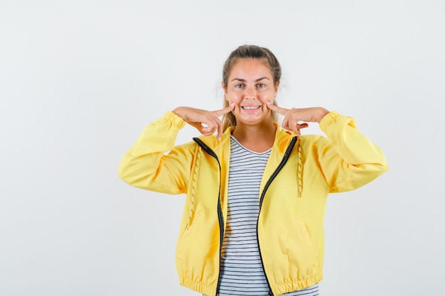 Junge Frau im T-Shirt, Jacke, die Finger auf Wangen hält und fröhlich schaut, Vorderansicht.