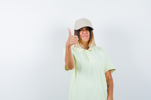 Junge Frau im T-Shirt, Kappe, die Daumen oben zeigt und fröhlich schaut, Vorderansicht.