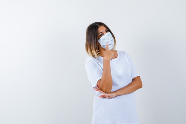 Junge Frau im T-Shirt, Maske zeigt nach oben und schaut fröhlich, Vorderansicht.