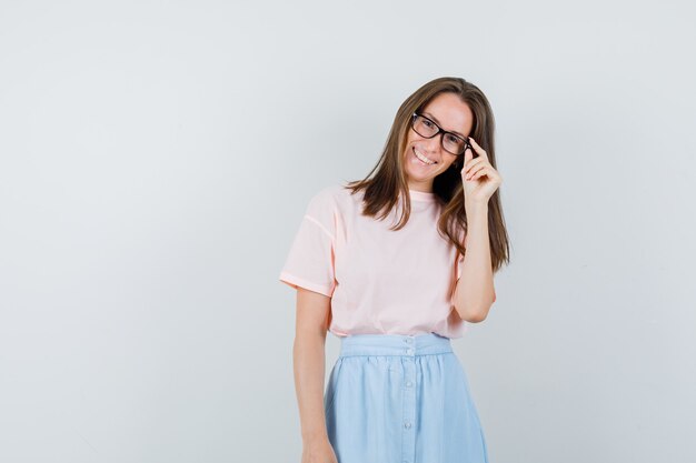 Junge Frau im T-Shirt, Rock, der Finger auf Brille hält und fröhlich, Vorderansicht schaut.