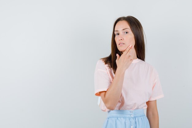 Junge Frau im T-Shirt, Rock, der Gesichtshaut mit den Fingern berührt, Vorderansicht.