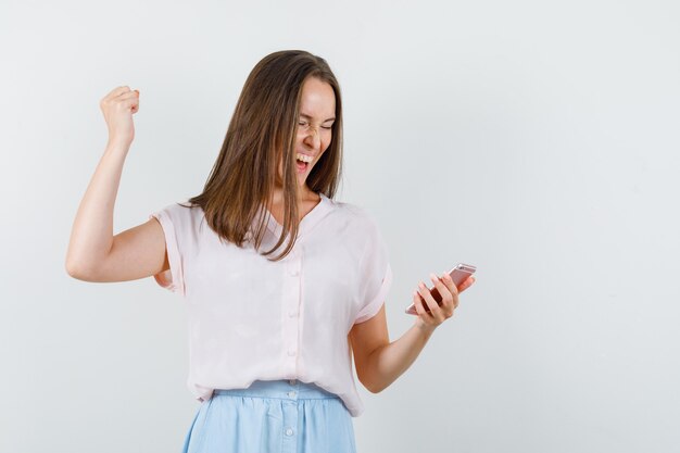 Junge Frau im T-Shirt, Rock, der Handy mit Siegergeste betrachtet und glücklich, Vorderansicht schaut.