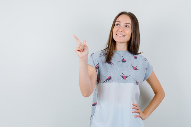 Junge Frau im T-Shirt zeigt nach oben und schaut hoffnungsvoll, Vorderansicht.