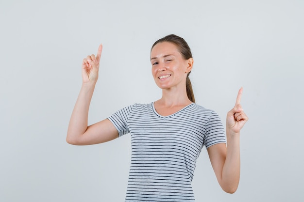 Junge Frau im T-Shirt zeigt nach oben und zwinkert Auge und schaut fröhlich, Vorderansicht.