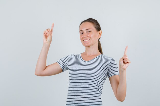 Junge Frau im T-Shirt zeigt nach oben und zwinkert Auge und schaut fröhlich, Vorderansicht.