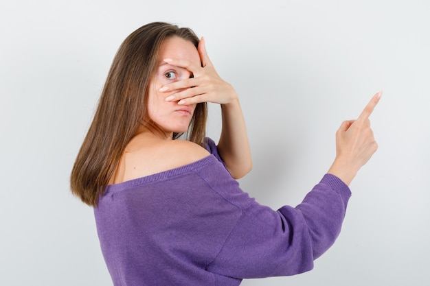 Junge Frau im violetten Hemd, das durch Finger schaut und weg zeigt.