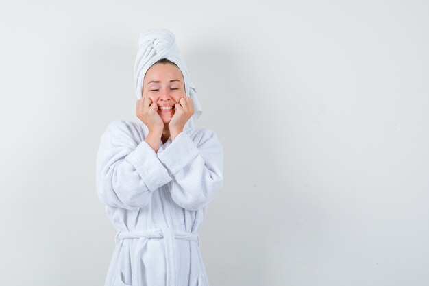 Junge Frau im weißen Bademantel, Handtuch, das Hände auf Wangen hält und fröhlich schaut, Vorderansicht.