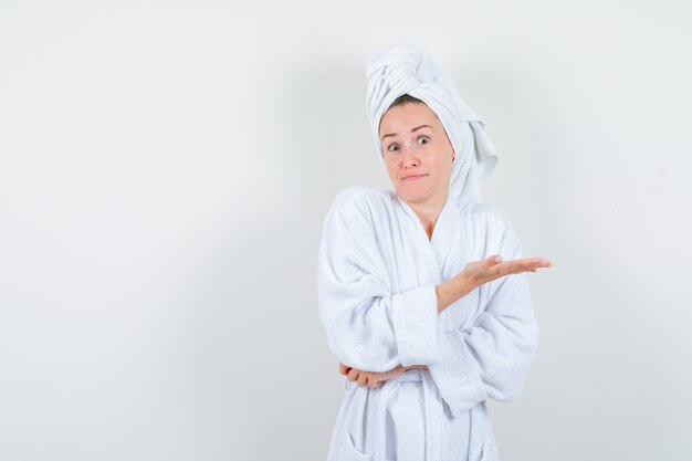 Junge Frau im weißen Bademantel, Handtuch, das Palme ausbreitet, um etwas zu zeigen und verwirrt zu sehen, Vorderansicht.