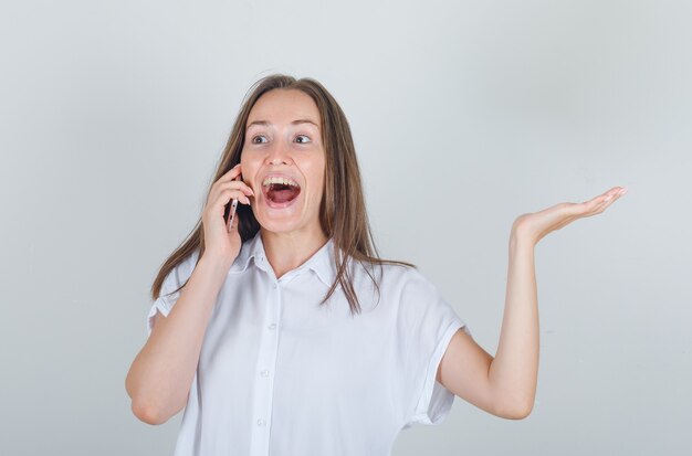 Junge Frau im weißen Hemd, das am Telefon mit Handgeste spricht und fröhlich schaut