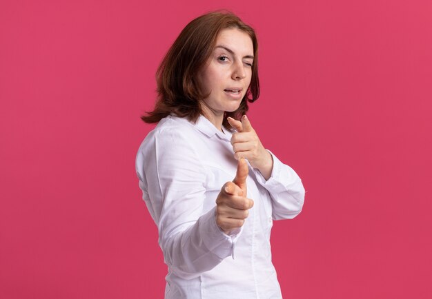 Junge Frau im weißen Hemd, das mit Zeigefingern an der Front zeigt, die zuversichtlich über rosa Wand stehend lächelt