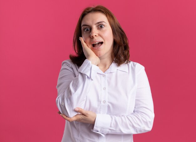 Junge Frau im weißen Hemd, das vorne erstaunt und überrascht mit Hand nahe Wange steht, die über rosa Wand steht