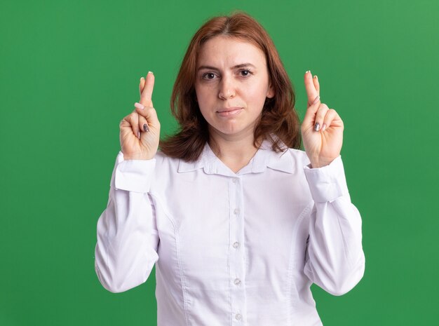 Junge Frau im weißen Hemd, das vorne mit ernstem Gesicht schaut, das Finger über grüner Wand kreuzt