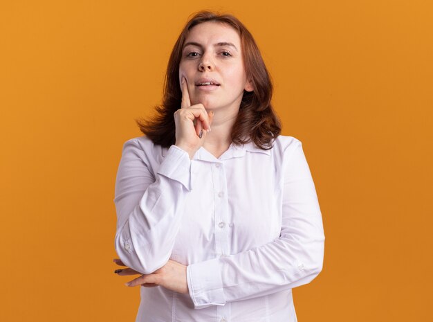Junge Frau im weißen Hemd, das vorne mit nachdenklichem Ausdruck betrachtet, der über orange Wand steht