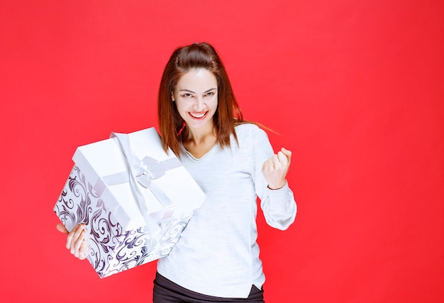 Junge Frau im weißen Hemd, die eine bedruckte Geschenkbox hält und ein positives Handzeichen zeigt