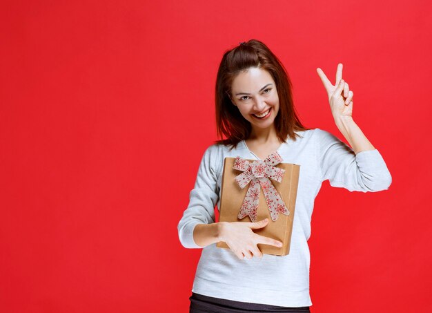 Junge Frau im weißen Hemd, die eine Geschenkbox aus Karton hält und ein positives Handzeichen zeigt