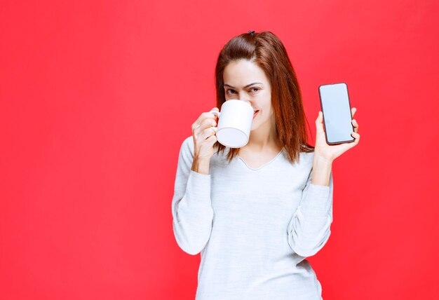 Junge Frau im weißen Hemd, die eine weiße Kaffeetasse und ein schwarzes Smartphone hält