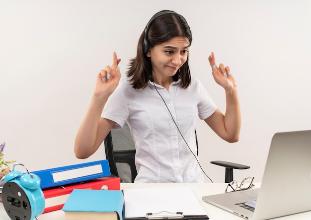 Junge Frau im weißen Hemd und in den Kopfhörern mit einem Mikrofon, das am Tisch mit Ordnern sitzt, die ihren Laptopbildschirm auf Videoanruf betrachten und wünschenswerten Wunsch kreuzen Finger über weiße Wand