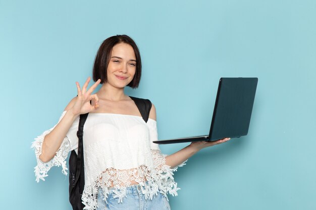 junge Frau im weißen Hemd und in der schwarzen Tasche, die Laptop hält und verwendet, der auf Blau lächelt