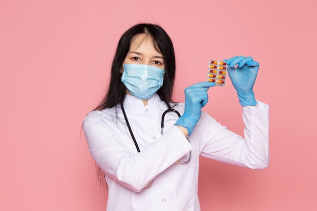 junge Frau im weißen medizinischen Anzug blaue Handschuhe blaue Schutzmaske, die mehrfarbige Pillen auf rosa hält