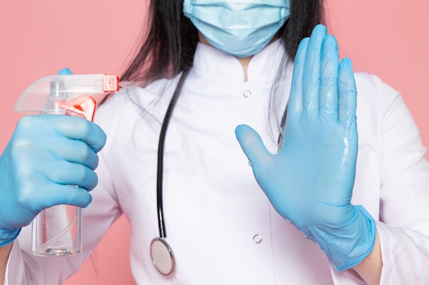 junge Frau im weißen medizinischen Anzug blaue Handschuhe blaue Schutzmaske mit Stethoskop, das desinfizierendes Spray auf rosa hält