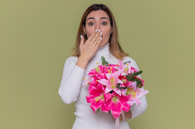 Junge Frau im weißen Rollkragenpullover, der Blumenstrauß hält, der an der Front erstaunt und überrascht bedeckt Mund mit Hand feiert internationalen Frauentag, der über grüner Wand steht