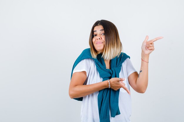 Junge Frau im weißen T-Shirt, die auf die Seite und nach unten zeigt und nachdenklich aussieht, Vorderansicht.