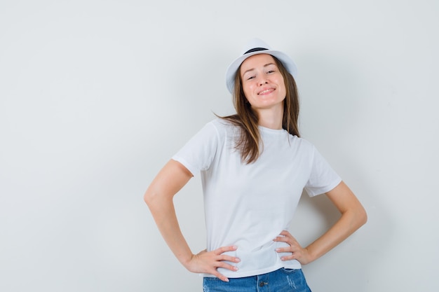 Junge Frau im weißen T-Shirt, in den kurzen Hosen, im Hut, der während des Stehens aufwirft und zuversichtlich schaut.