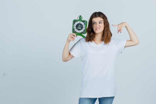 Junge Frau im weißen T-Shirt, Jeans, die auf Uhr zeigen, Stirnrunzeln und unruhig aussehen, Vorderansicht.