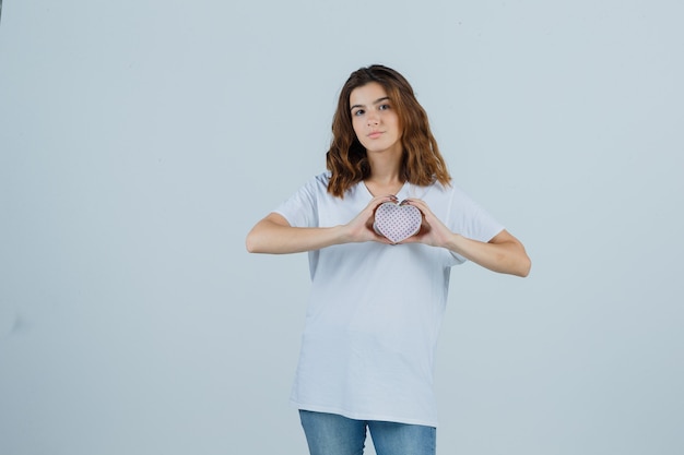 Junge Frau im weißen T-Shirt, Jeans, die Geschenkbox hält und selbstbewusst, Vorderansicht schaut.