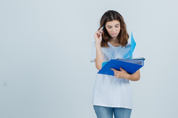 Junge Frau im weißen T-Shirt, Jeans, die Ordner und Stift halten und nachdenklich schauen, Vorderansicht.