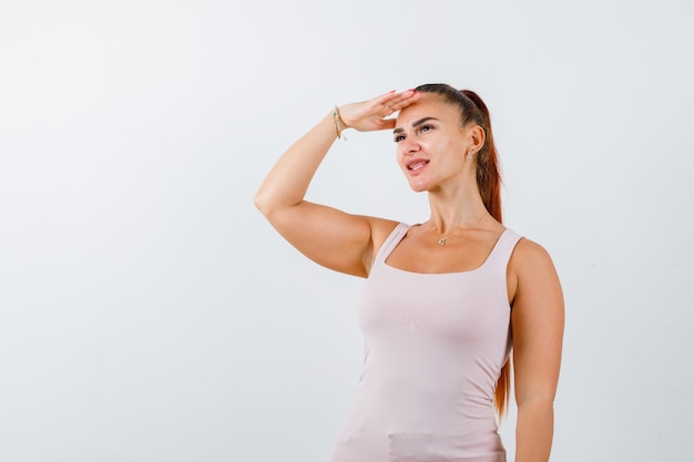 Junge Frau im weißen Trägershirt, das Hand auf Kopf hält, um klar zu sehen und hübsch, Vorderansicht aussehend.