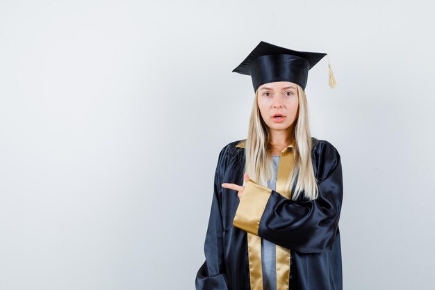 Junge Frau in Absolventenuniform, die auf die linke Seite zeigt und verwirrt aussieht