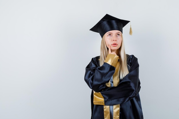 Kostenloses Foto junge frau in absolventenuniform, die in denkender pose steht und zögerlich aussieht