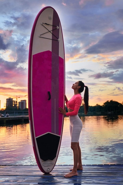 Junge Frau in Activewear steht mit Sup-Board am Pier