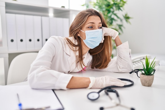 Junge Frau in Arztuniform und medizinischer Maske sitzt müde am Tisch in der Klinik