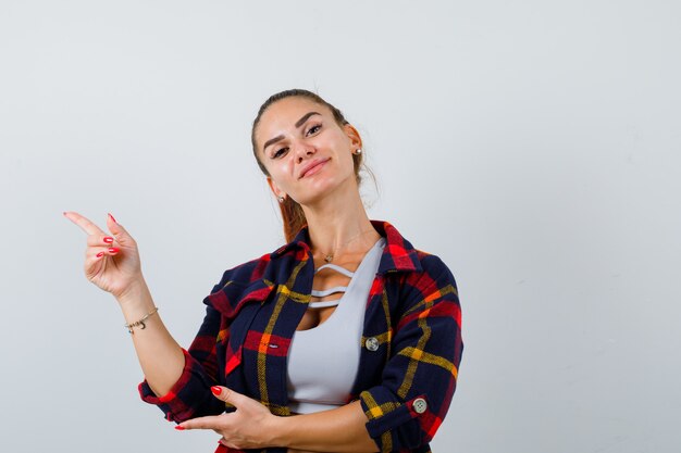 Junge Frau in bauchfreiem Top, kariertem Hemd, das auf die obere linke Ecke zeigt und selbstbewusst aussieht, Vorderansicht.